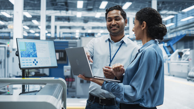 Indian Male Engineer And Hispanic Female Supervisor Using Laptop And Talking At Electronics Factory. Man Using Soldering Jet Printer, Explaining Process Behind Production Of Circuit Boards.
