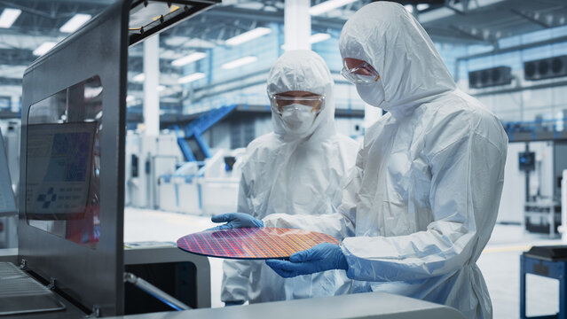 Semiconductor Production Process At Electronics Factory: Two Technicians In Sterile Suits Taking Out Silicon Wafer From Soldering Jet Printer And Inspecting It. Engineers Making Hardware For Computers