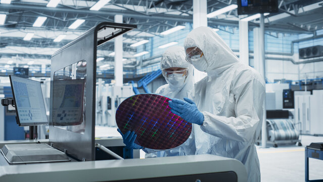 Semiconductor Production At Electronics Manufacturing Facility: Two Specialists In Sterile Suits Taking Out Silicon Wafer From Soldering Jet Printer And Checking It. Engineers Making Computer Hardware