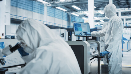 Scientists In Sterile Suites In Electronics Factory Cleanroom: Engineer Using Microscope And Desktop Computer To Inspect Microchip. His Colleague Using Soldering Jet Printer To Print Motherboards.