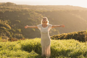 A woman in a light dress smiles joyfully with open arms, surrounded by lush greenery under a soft...