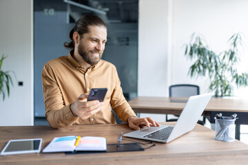 Fototapeta premium A modern office worker multitasking using a phone and laptop at a wooden table in an office. The scene conveys productivity and professional engagement with technology in a corporate environment.