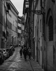 Naklejka premium A narrow cobbled street in Florence framed by elegant historic buildings, with pedestrians strolling through. The old-world charm is evident in the street lamps.