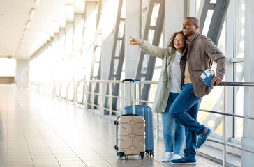 Joyful African American Couple Standing With Luggage At Airport And Pointing Away, Happy Young...