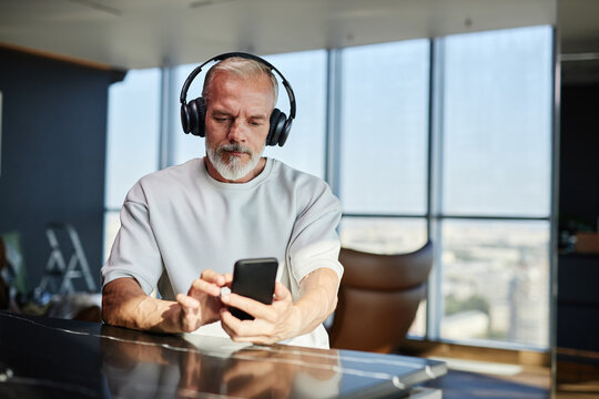 Medium shot of mature man with well groomed white beard listening to music in headphones while using mobile phone at table in contemporary hotel room, copy space