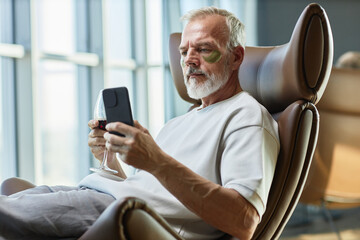 Medium shot of mature businessman taking care of face skin with eye patches, while text messaging on mobile phone over glass of red wine in comfy armchair at home