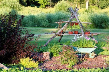 A picturesque garden scene featuring a rustic wooden trellis with flowerpots, an old wheelbarrow...