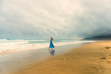 A woman in a flowing blue dress running barefoot on the sandy Playa de Cofete beach in Fuerteventura, Canary Islands. The dramatic cloudy sky, golden sand, and ocean waves 