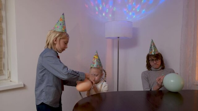 Three young children wearing party hats inflate colorful balloons in slow motion, creating a joyful and festive atmosphere