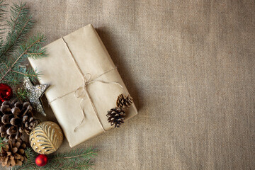 A gift wrapped in paper is placed on a burlap background, surrounded by Christmas decorations. Copy space. Vintage style.