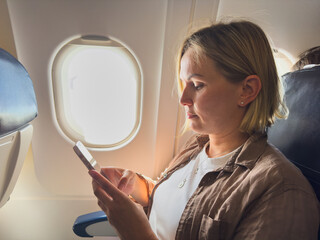 Fototapeta premium Passenger woman is flying in plane. Girl using phone sitting by airplane window. Traveling female with smartphone inside plane enjoying flight. Woman using wifi connection on board. Traveling girl
