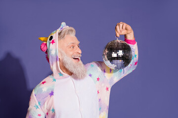 Joyful mature man in colorful unicorn pajamas holding disco ball against purple backdrop