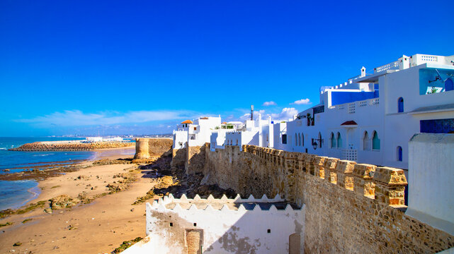 White buildings with colorful windows, typical of the architecture of Asilah, Morocco, with blue and green details. Under a bright sky, the atmosphere evokes the peaceful charm of this coastal town.