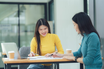 Fototapeta premium Collaborative Teamwork: Two young Asian women engaged in a productive meeting, reviewing documents and ideas together in a modern workspace. 
