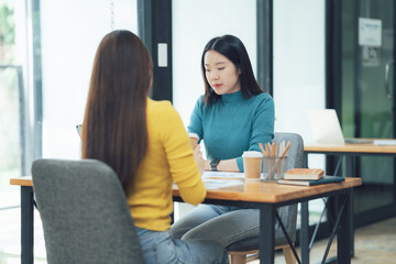 Fototapeta premium Collaborative Meeting: Two young Asian women engage in a focused discussion, leaning in attentively during a collaborative meeting at a modern office desk.