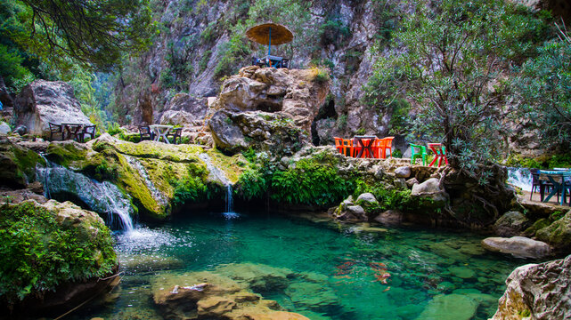 
A natural turquoise pool in Akchour, Chefchaouen, surrounded by lush green rocks. Colorful tables and a rustic parasol create a relaxing setting in the heart of nature.