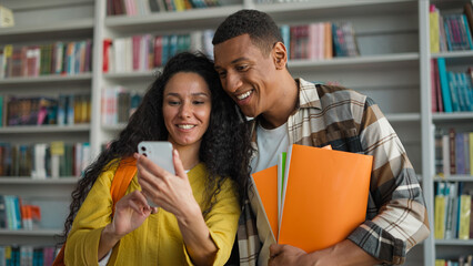 Two people students African American man Indian Hispanic woman holding mobile phone smartphone social media online laughing girl guy library university high school classmates friends studying learning