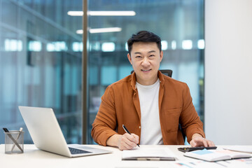 Asian man, mature businessman working at his desk, using laptop and writing notes.