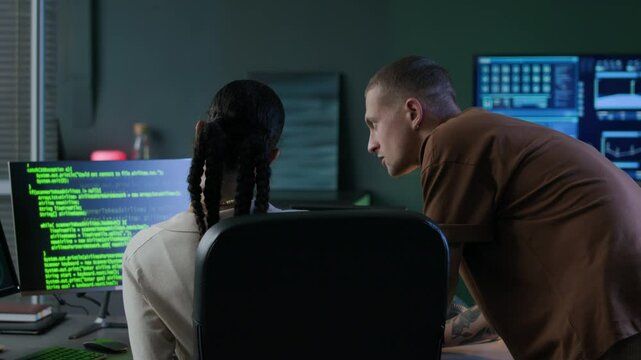 Back view shot of young male Caucasian programmer checking program code of his junior female biracial colleague and complimenting on her correct work with high five in modern office at night