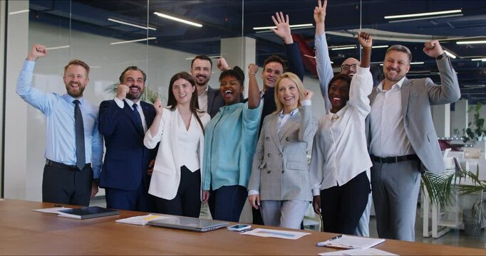 Business team having fun after a successful meeting. Group of happy, overjoyed multiracial people standing in the office, smiling, raising hands up, and cheering