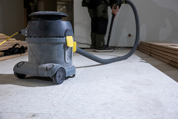 A vacuum cleaner and worker in protective gear prepare for floor installation. Wooden planks are stacked along the wall, indicating ongoing work indoors.