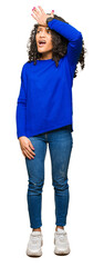 Young beautiful woman with curly hair wearing winter sweater surprised with hand on head for...