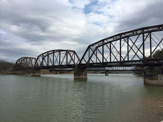 Fototapeta premium A rusty truss bridge spans a calm river under a cloudy sky. Its intricate metal framework reflects in the water, showcasing historic charm and timeless industrial engineering.