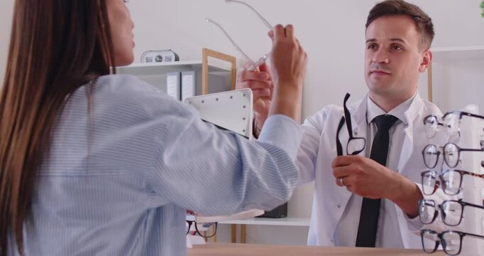 Young woman is visiting optometrist and trying to pick up new eyeglasses. Male ophthalmologist, optician doctor showing different kind of glasses with various frames, smiling and nodding approvingly.
