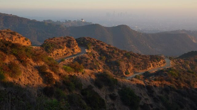180-degree dynamic panorama highlighting Runyon Canyon's hiking terraces, Griffith Observatory, hillside homes, and the hazy downtown LA skyline at golden hour.