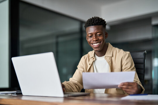 Happy busy young African business man checking documents working at laptop in office. Smiling businessman accountant company employee reading law paper corporate file finances invoice report.