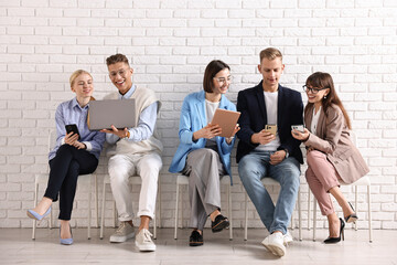 Fototapeta premium Group of people using different gadgets on chairs near white brick wall indoors. Modern technology