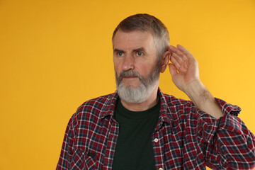 Senior man showing hand to ear gesture on orange background