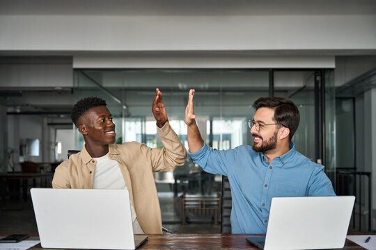Two happy diverse young professional male employees team giving high five at work. African and Hispanic business men buddies celebrating good corporate teamwork results, startup success together.