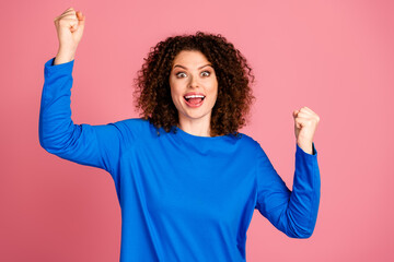 Excited young woman with curly hair in blue sweater celebrating against pink background