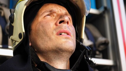 Young tired fireman in protective uniform against the background of a fire engine. Portrait of male firefighter with dirty face near fire truck. Concept of saving lives and heroic profession. Close up