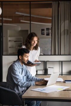 Indian and Latina teammates working on joint project, male mentor explain task details to female apprentice, looking at laptop screen, provide new details, intern take notes, learns corporate software