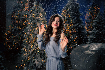 Fototapeta premium Woman smiles in a winter forest with snowfall. Evergreen trees and holiday lights, festive atmosphere for Christmas and New Year celebrations.