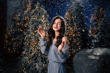 Fototapeta premium Woman smiles in a winter forest with snowfall. Evergreen trees and holiday lights, festive atmosphere for Christmas and New Year celebrations.
