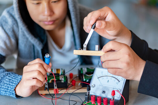 Children are studying electrical circuit systems by experimenting with electrical connections using a small electric meter and a small flashlight bulb in physics science class, soft focus.