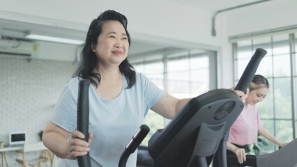  Asian senior woman exercising on elliptical trainer with daughter in living room at home.