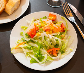 Plate contains portion of salad from fresh China cabbage, grated carrot, corn, tomato complemented by small glass of wine.