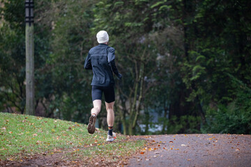 Obraz premium A man in a gray sweatshirt and black shorts jogs along a damp park trail in Portland, OR. Surrounded by greenery and fresh air, the peaceful setting makes for a healthy outdoor activity