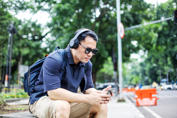 Obraz premium Tourist Guy Wear Headphone Hearing Music Sitting on Sidewalk While Using Smartphone