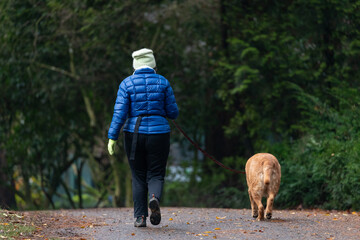 A woman in a blue puffer jacket and white beanie walks her golden retriever along a tranquil park...
