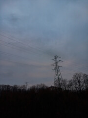 Silhouette of trees without leaves as it is winter time behind which the roof of some house is being monitored and above all that a power line with cables in cloudy weather