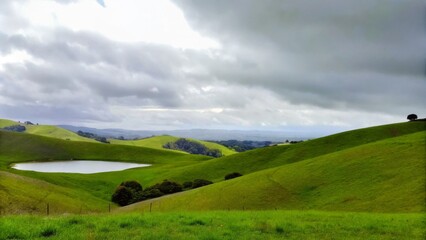 Fototapeta premium Rolling Green Hills and a Lone Reflective Pond Under a Moody Sky for Scenic Photography 