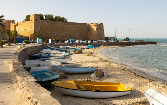 Tranquil scenic view of Hammamet coastal promenade with local fishing boats lined up on shore against backdrop of imposing ancient fortified Kasbah, guarding city under clear sky of Tunisia.