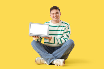 Young man with blank laptop sitting on yellow background