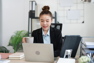 Fototapeta premium Happy Asian businesswoman working and checking work with laptop computer while having a comfortable cup of coffee at office.