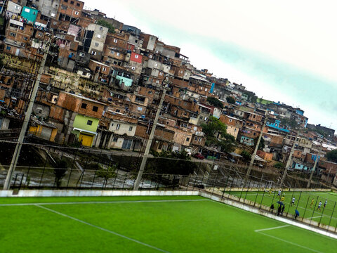 Campo de Futebol de V&aacute;rzea com Grama Sint&eacute;tica e Comunidade ao Fundo